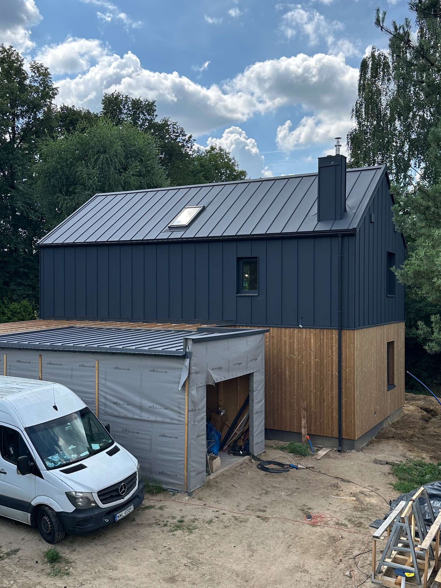Dwukondygnacyjny dom szkieletowy w technologii Steico z ciemnoszarą blachą na dachu i drewnianą fasadą, częściowo osłonięty folią, z białym busem na podjeździe.