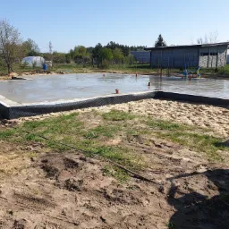 Wylany fundament z wystającymi prętami zbrojeniowymi, zabezpieczony czarną folią izolacyjną, otoczony piaskiem i ziemią, w tle budynek gospodarczy i drzewa.