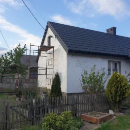 Dom jednorodzinny z szarą dach&oacute;wką, częściowo ocieplony białym styropianem, z widocznym rusztowaniem przy ścianie szczytowej i drewnianym płotem w ogrodzie.