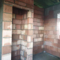 Wnętrze domu w stanie surowym: ściany z czerwonej ceramiki, otwór drzwiowy z ościeżnicą z tego samego materiału, widoczne zielone elementy konstrukcji dachu, surowa podłoga.