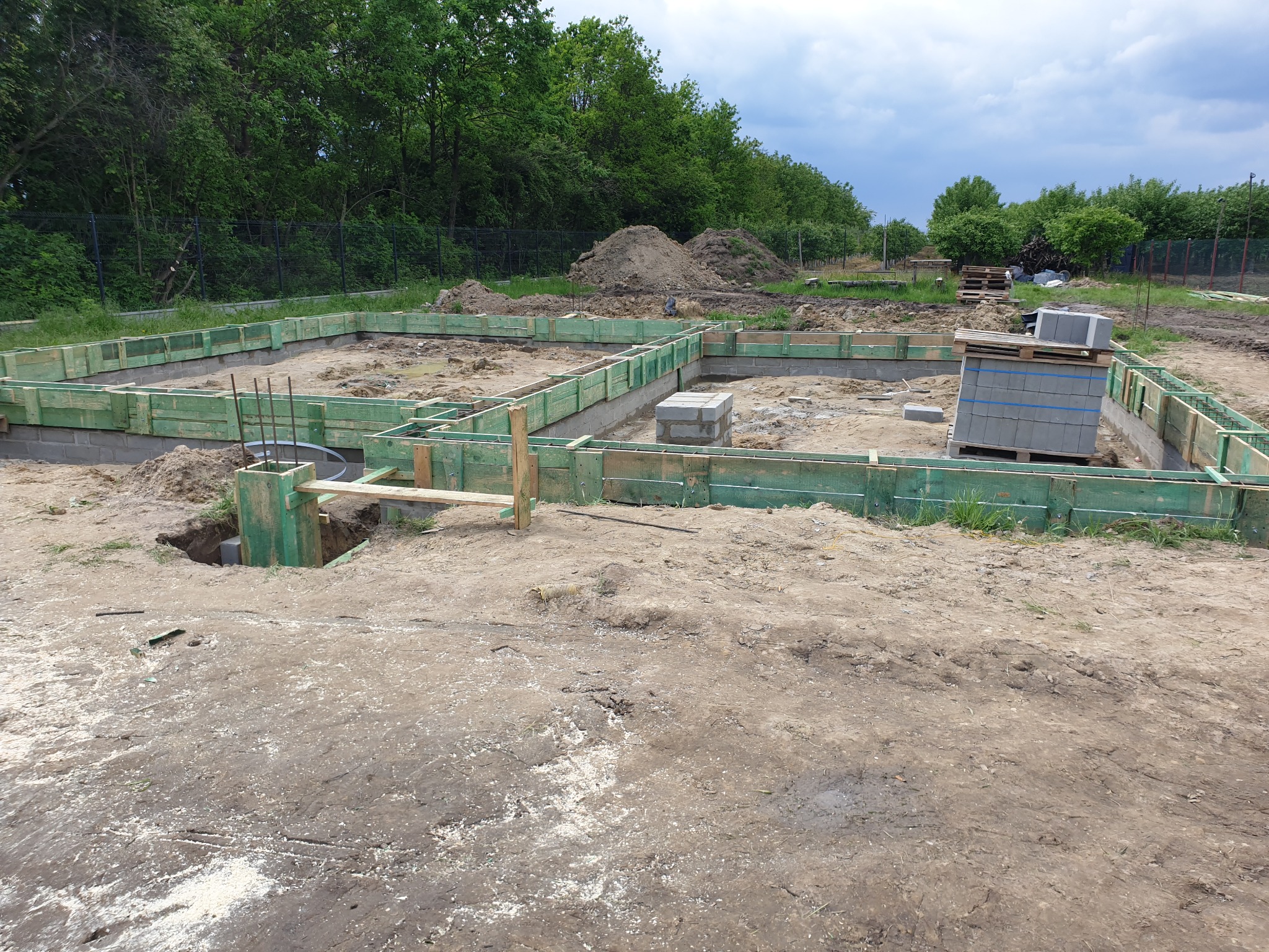 Przygotowany szalunek i częściowo wylane fundamenty pod budynek, widoczne zbrojenia, bloczki betonowe na palecie i wykopany grunt, w tle zieleń drzew i pochmurne niebo.