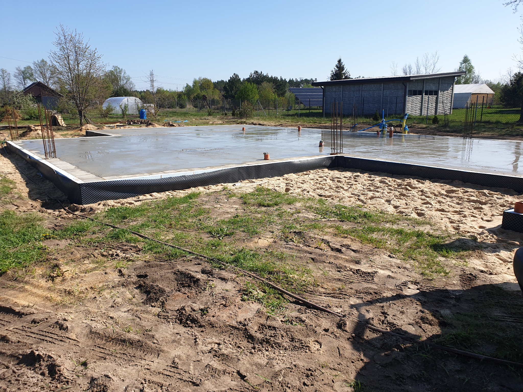 Wylany fundament z wystającymi prętami zbrojeniowymi, zabezpieczony czarną folią izolacyjną, otoczony piaskiem i ziemią, w tle budynek gospodarczy i drzewa.