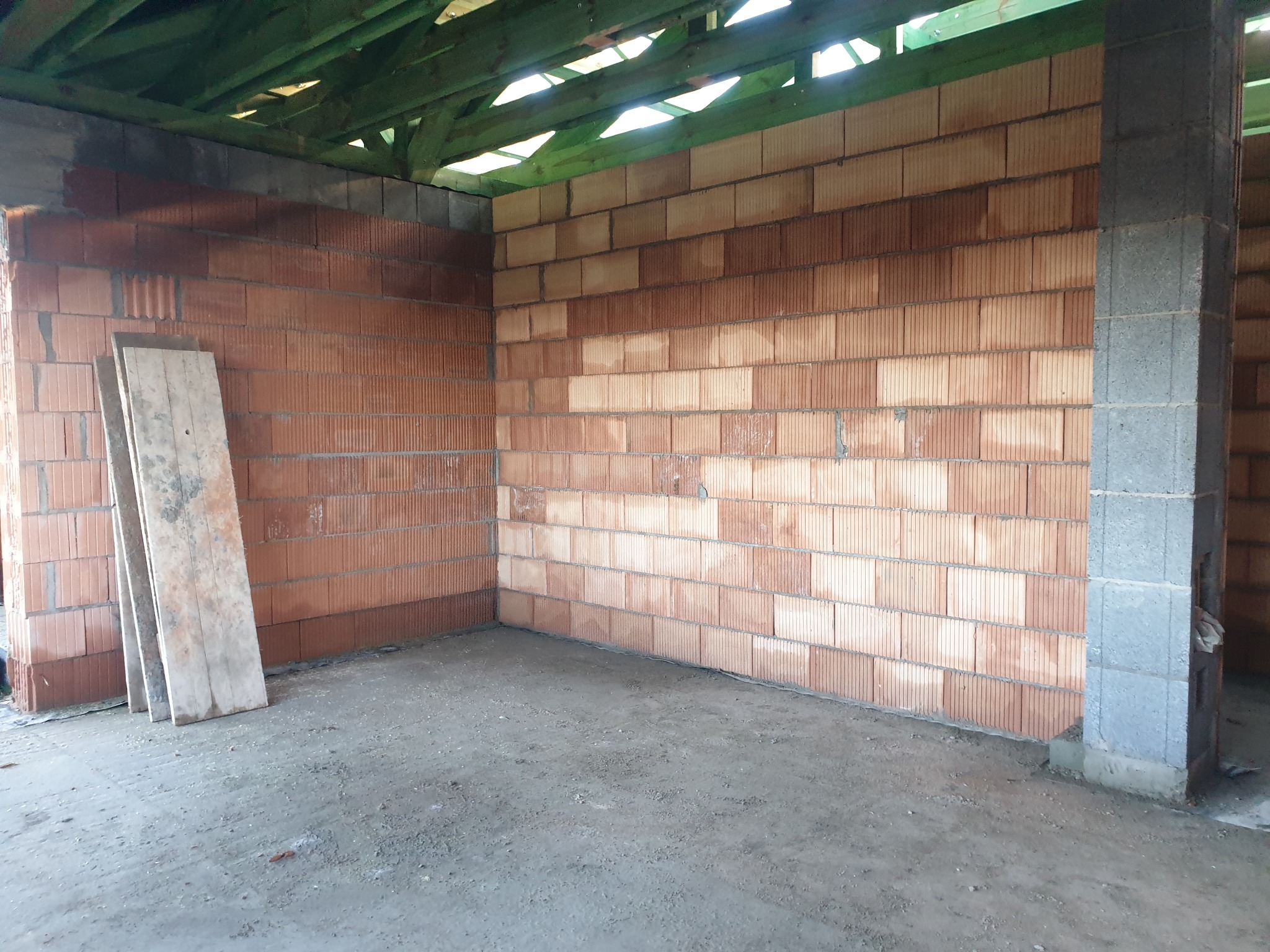 Wnętrze w trakcie budowy z widocznymi ścianami z bloczków ceramicznych, surowym betonem na podłodze i elementami drewnianej konstrukcji dachu, trzy deski oparte o ścianę w lewym dolnym rogu.