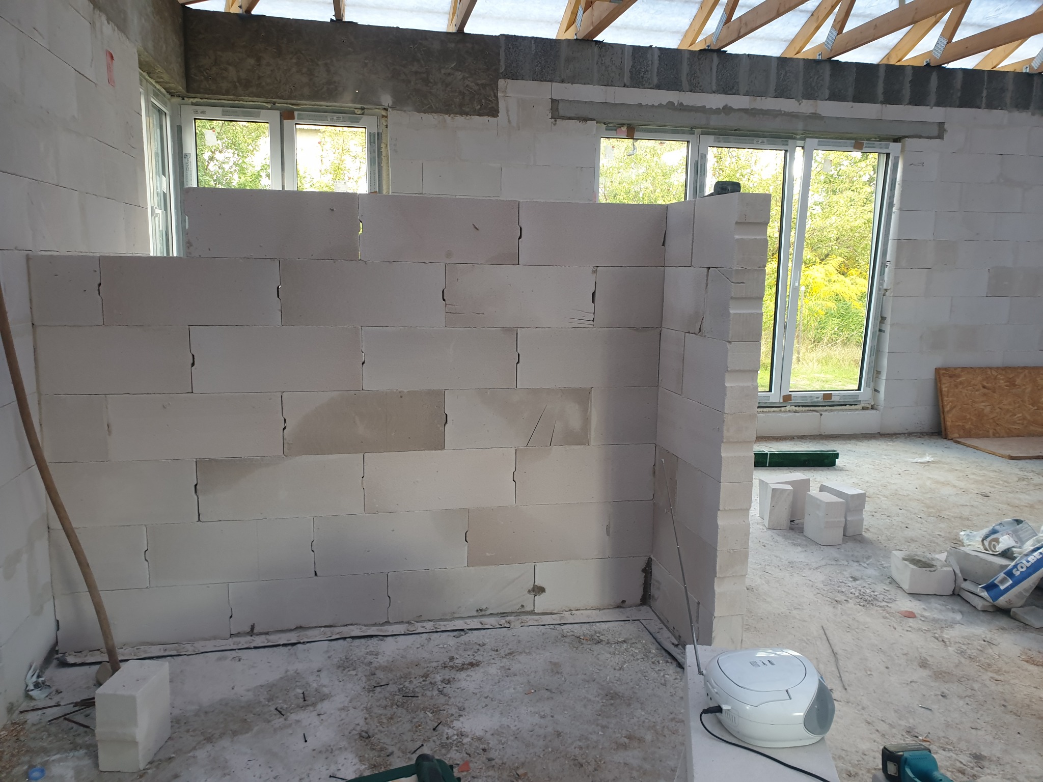 Wnętrze w trakcie wznoszenia ścian działowych z bloczków betonowych, widoczne okna, surowy strop i rozłożone narzędzia oraz radio.