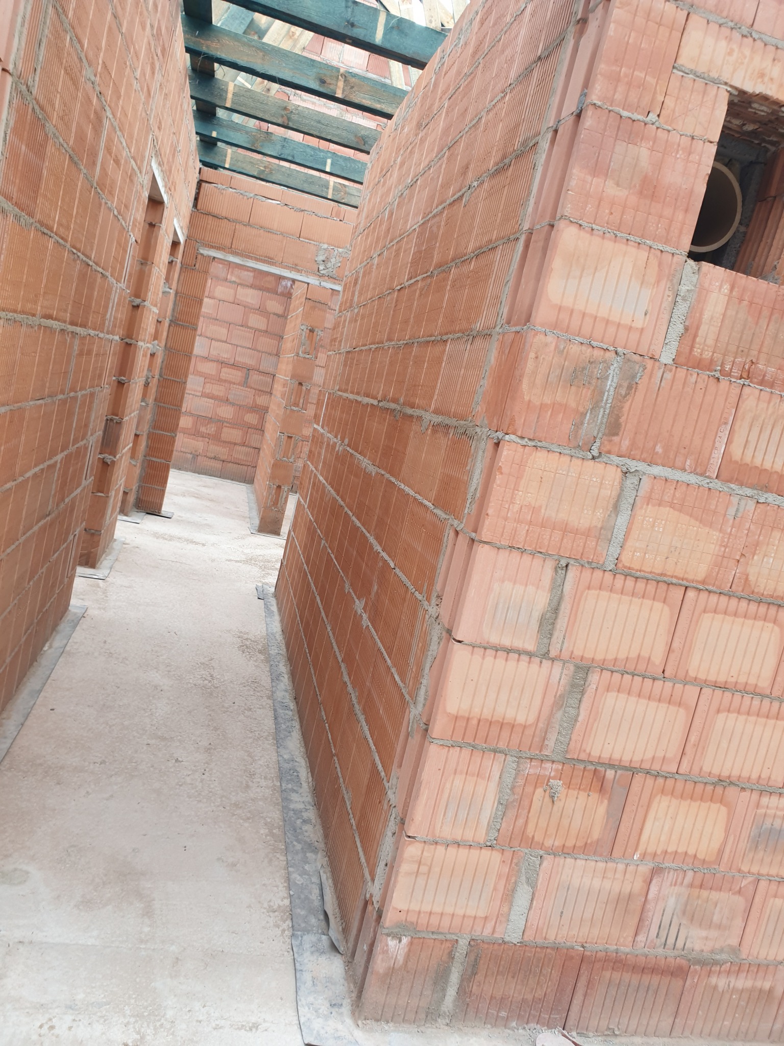 Wnętrze budynku w trakcie budowy, widoczne ściany z czerwonej cegły ceramicznej, surowy beton na podłodze i drewniana konstrukcja dachu w tle.