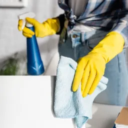 Osoba w żółtych rękawicach przeciera wilgotną, błękitną ściereczką białą powierzchnię laptopa, obok stoi niebieska butelka ze spryskiwaczem, w tle widoczna roślina doniczkowa.
