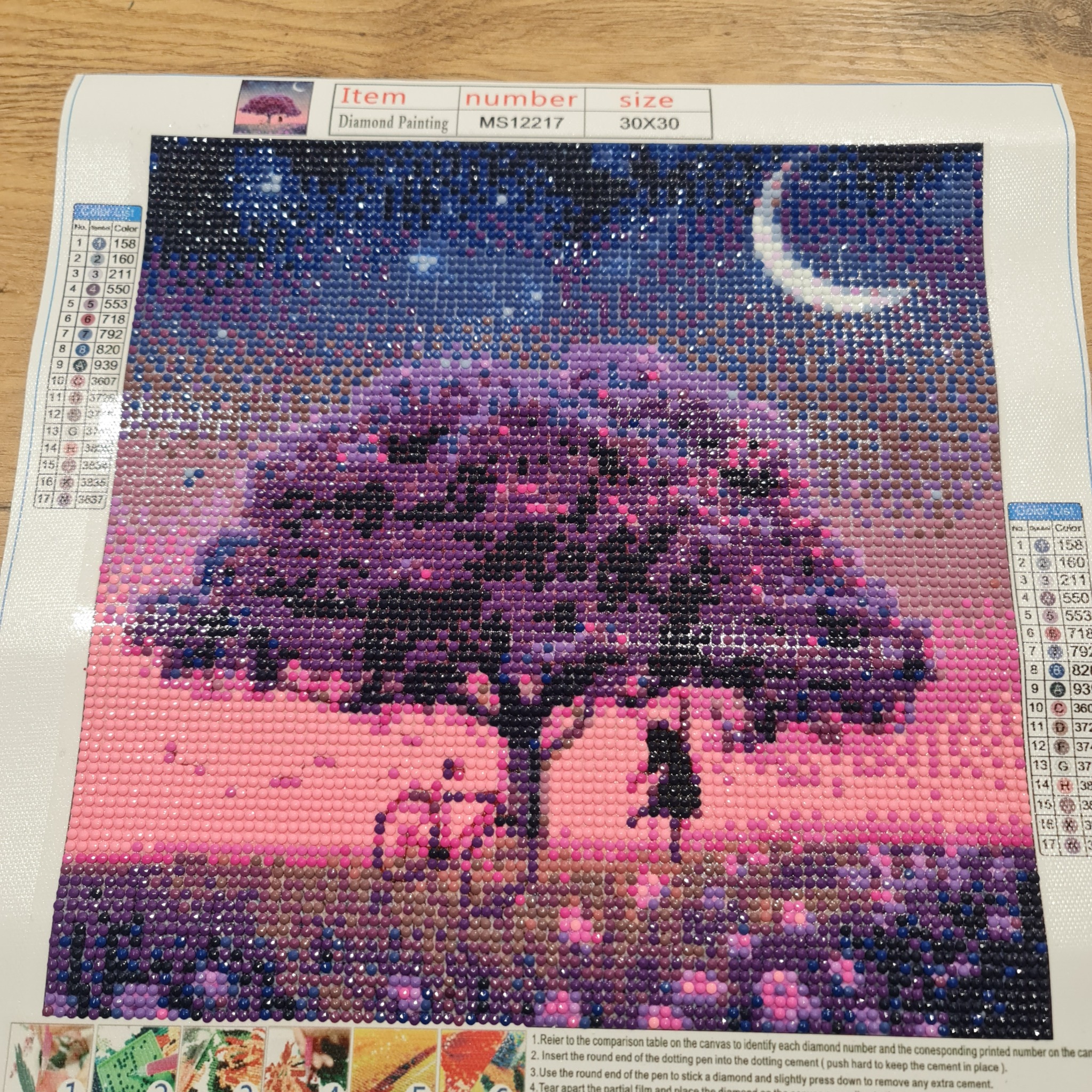 Diamond painting przedstawiający nocny krajobraz z drzewem, rowerem i sylwetką osoby pod księżycem, wykonany z drobnych, błyszczących diamencików o różnych kolorach, widoczna legenda kolorów...