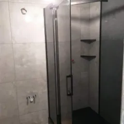 Nowoczesna kabina prysznicowa z czarnymi profilami i szklanymi drzwiami, zamontowana w łazience z szarymi płytkami wielkoformatowymi i drewnopodobną podłogą, widoczne niedokończone podłączenie...