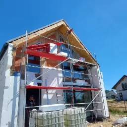 Budowa domu jednorodzinnego z białych bloczków, widoczne rusztowania, okna i przygotowywana elewacja na tle błękitnego nieba.