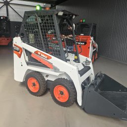 MAR MAX - Biała ładowarka Bobcat S70 z pomarańczowymi felgami stoi w magazynie z szarymi ścianami, w tle widoczne inne maszyny budowlane.