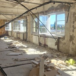 MAR MAX - Wnętrze budynku w trakcie rozbiórki, widoczne fragmenty ścian, gruz, odsłonięte instalacje oraz okna z widokiem na okolicę.