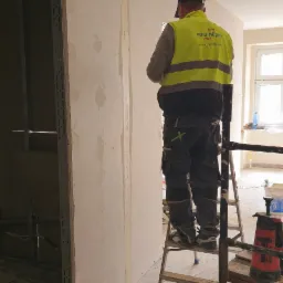 Pracownik w odblaskowej kamizelce na drabinie, szpachluje ścianę z płyt karton-gips. Widoczne profile i narzędzia. Ściana częściowo wykończona.
