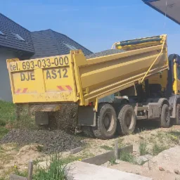 Żółta wywrotka wysypuje żwir na placu budowy domu jednorodzinnego w słoneczny dzień.