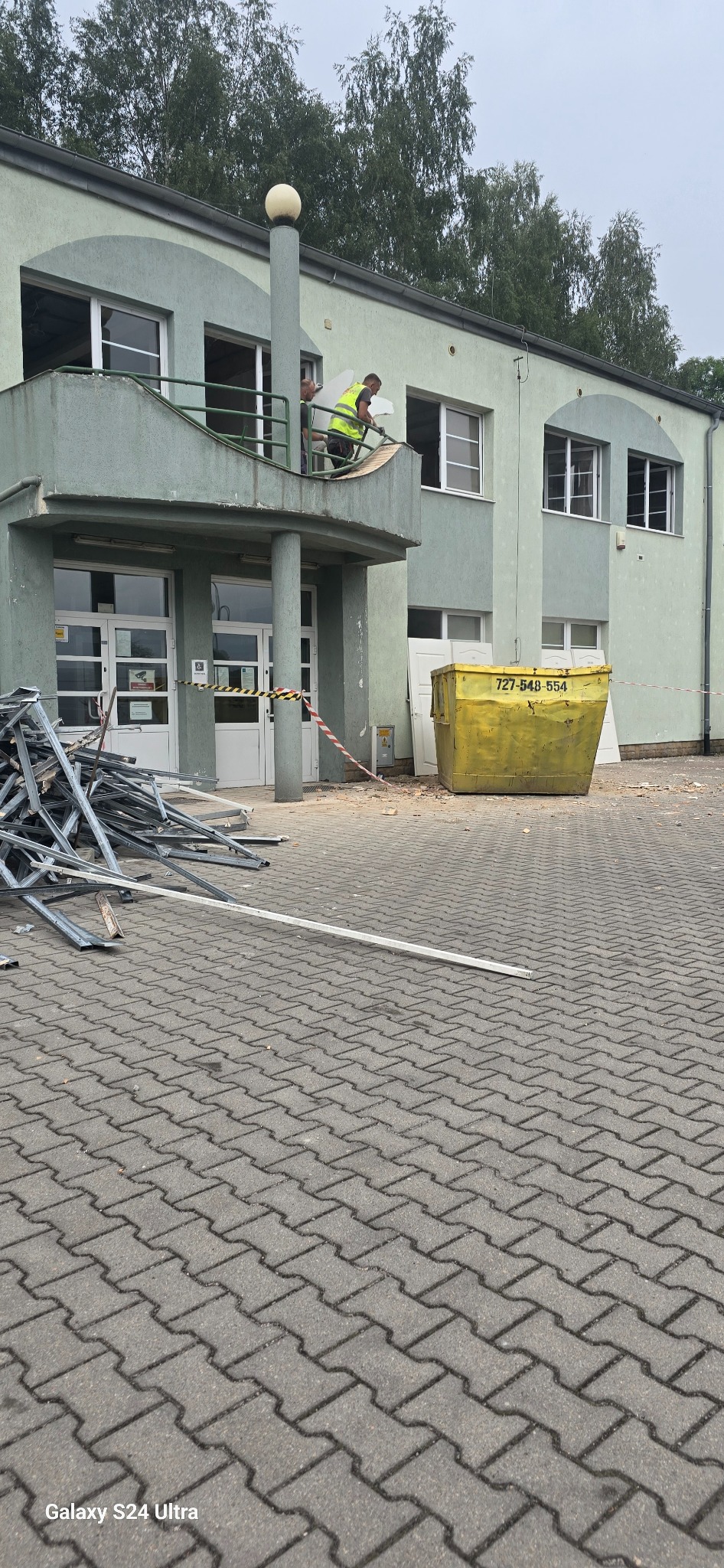 Dwóch pracowników w kamizelkach odblaskowych demontuje elementy budynku z balkonu, widoczny kontener na gruz, pozostałości konstrukcji na brukowanym placu przed budynkiem.