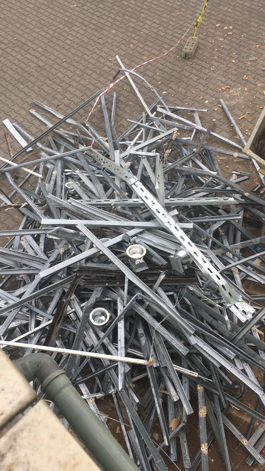 Sterta pociętych, metalowych profili konstrukcyjnych po demontażu, widoczne fragmenty instalacji elektrycznej i rury kanalizacyjnej, ujęcie z góry.