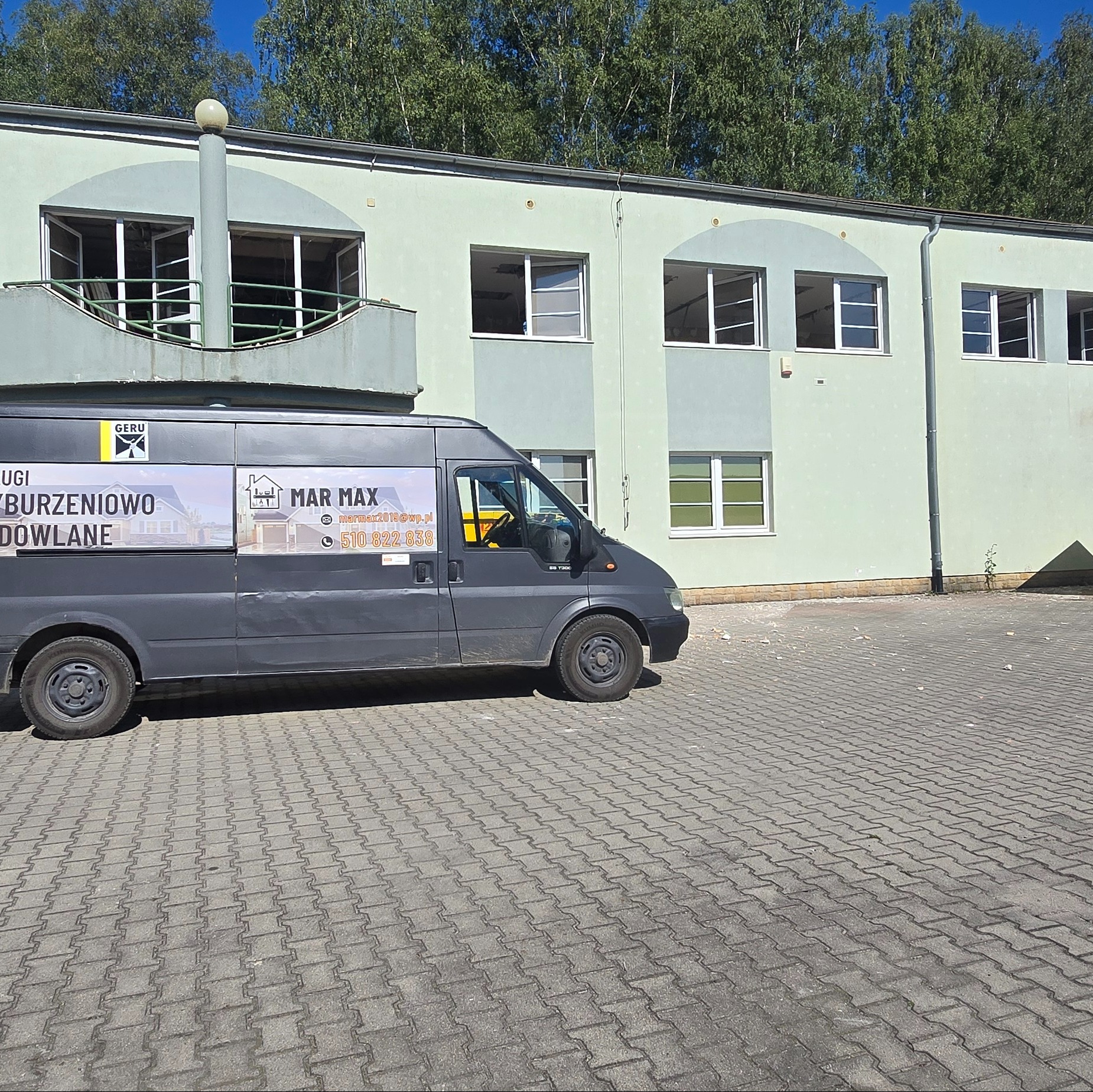 Szary bus firmy budowlanej MAR MAX z reklamą usług wyburzeniowych zaparkowany przed budynkiem z odpadami budowlanymi na kostce brukowej.