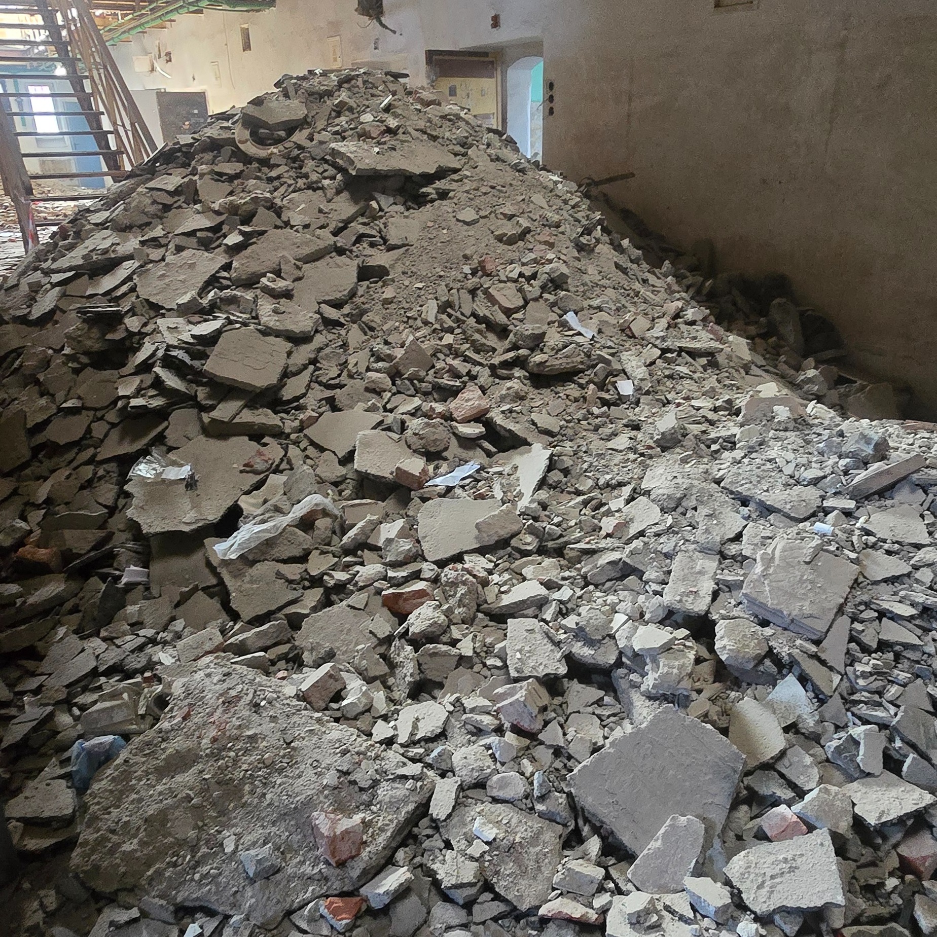 Sterta gruzu po wyburzeniu, widoczne fragmenty cegieł i betonu, w tle schody i drzwi do innego pomieszczenia.