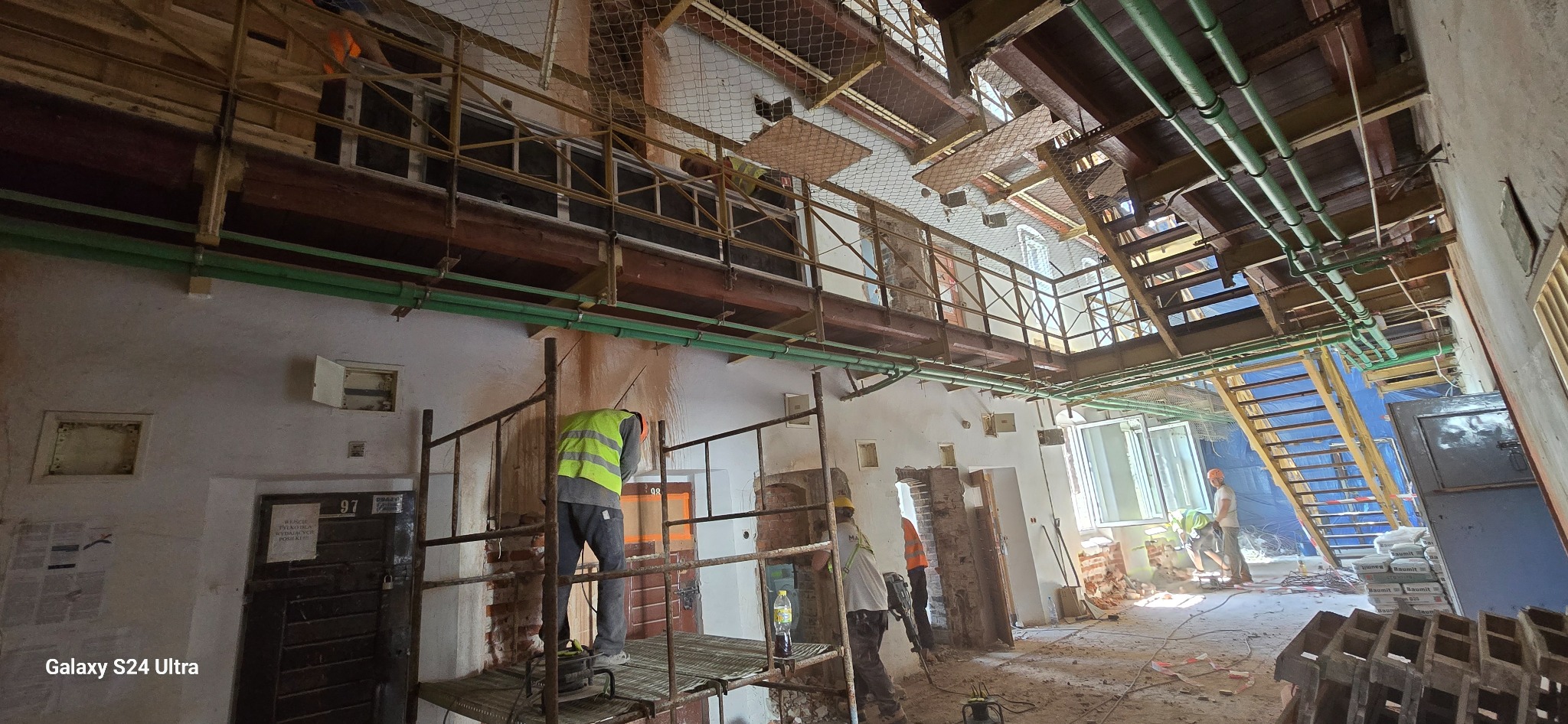 Wnętrze budynku w trakcie prac rozbiórkowych: widoczne rusztowania, pracownicy w kamizelkach odblaskowych, odsłonięte cegły po wyburzeniach, instalacje rurowe pod sufitem, worki z materiałami...