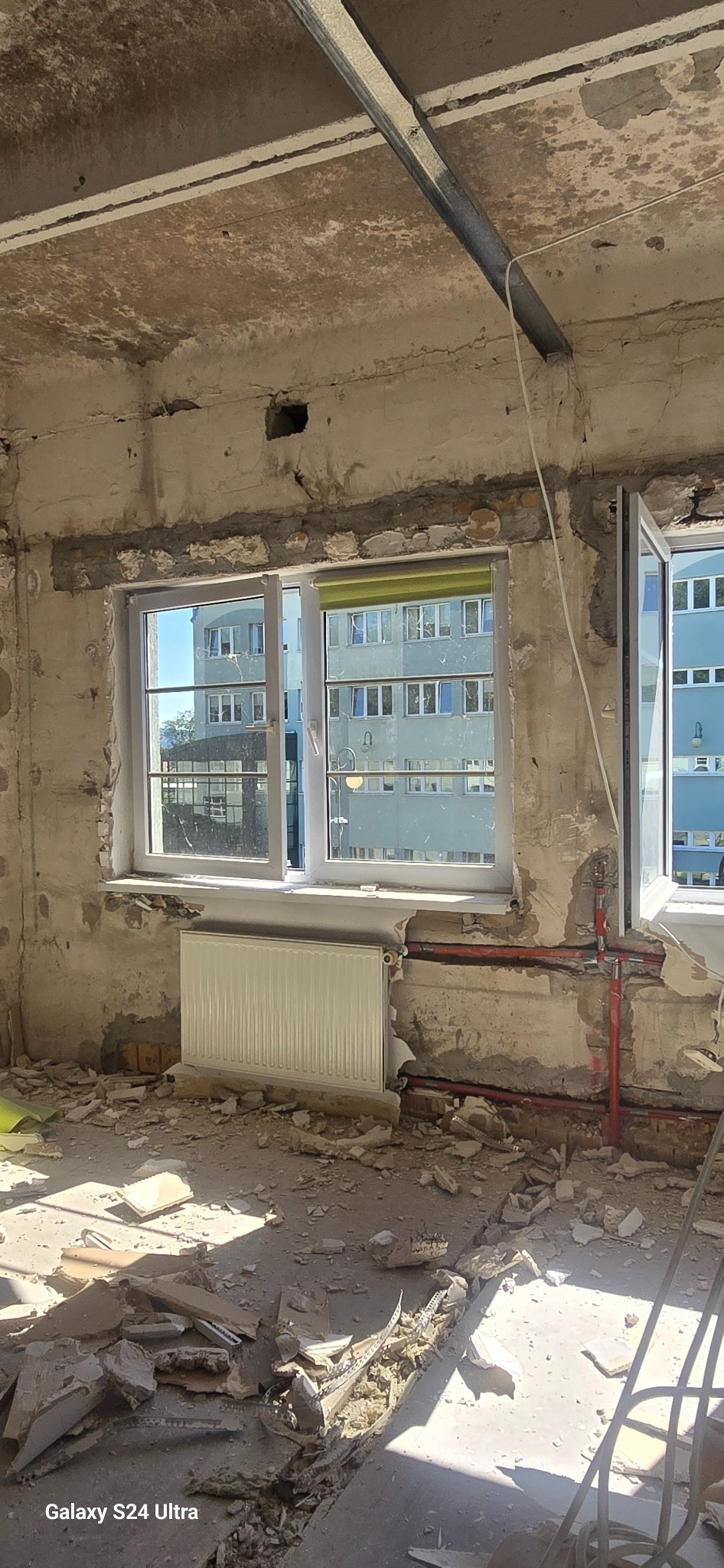 Wnętrze mieszkania w trakcie remontu: odsłonięte ściany, gruz na podłodze, grzejnik pod oknem, metalowy profil pod sufitem, widoczne instalacje grzewcze.