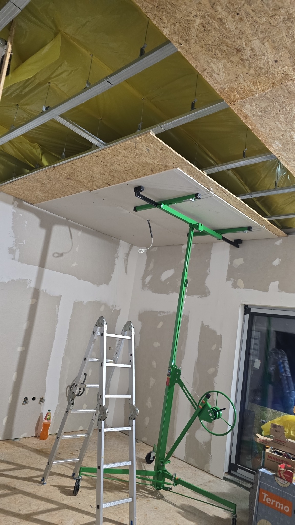 Montaż płyty gipsowo-kartonowej na suficie za pomocą podnośnika, widoczna aluminiowa drabina i częściowo przygotowane ściany do malowania.