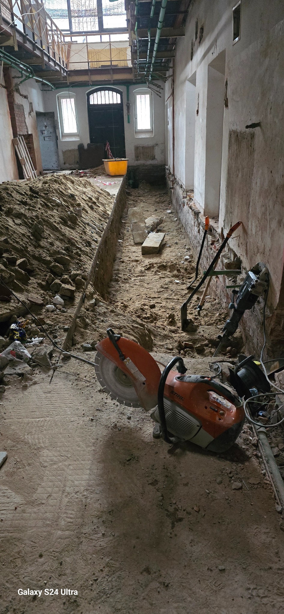 Wnętrze w trakcie prac wyburzeniowych, widoczny wykop w ziemi, narzędzia takie jak piła do betonu Stihl, młot pneumatyczny, łopaty oraz gruz i piasek.
