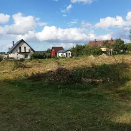 Teren po wycince drzew i krzewów z widocznymi budynkami mieszkalnymi w tle, błękitne niebo z chmurami.