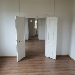 Wnętrze pokoju z drewnianą podłogą i otwartymi białymi drzwiami, widok na kolejne pomieszczenia w perspektywie, surowy drewniany sufit.