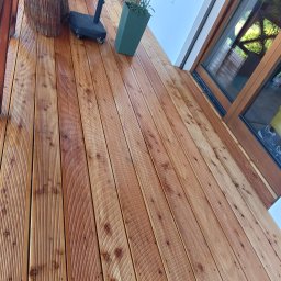 Prace remontowo-wykończeniowe - Świeżo ułożona, impregnowana deska tarasowa z widocznymi słojami, donicą z rośliną i fragmentem mebla ogrodowego.