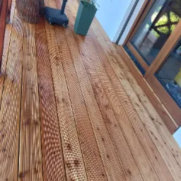 Świeżo ułożona, impregnowana deska tarasowa z widocznymi słojami, donicą z rośliną i fragmentem mebla ogrodowego.