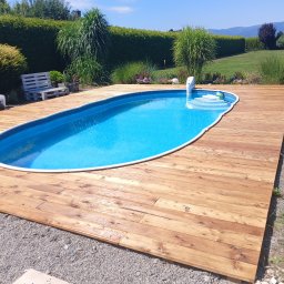 Prace remontowo-wykończeniowe - Niebieski basen otoczony drewnianym tarasem, z widokiem na zielony trawnik, ozdobne krzewy i góry w tle.