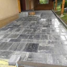 Prace remontowo-wykończeniowe - Ułożona kostka brukowa na tarasie przylegającym do budynku z drewnianą konstrukcją zadaszenia, widoczne niedokończone obrzeża i zapas kostki.