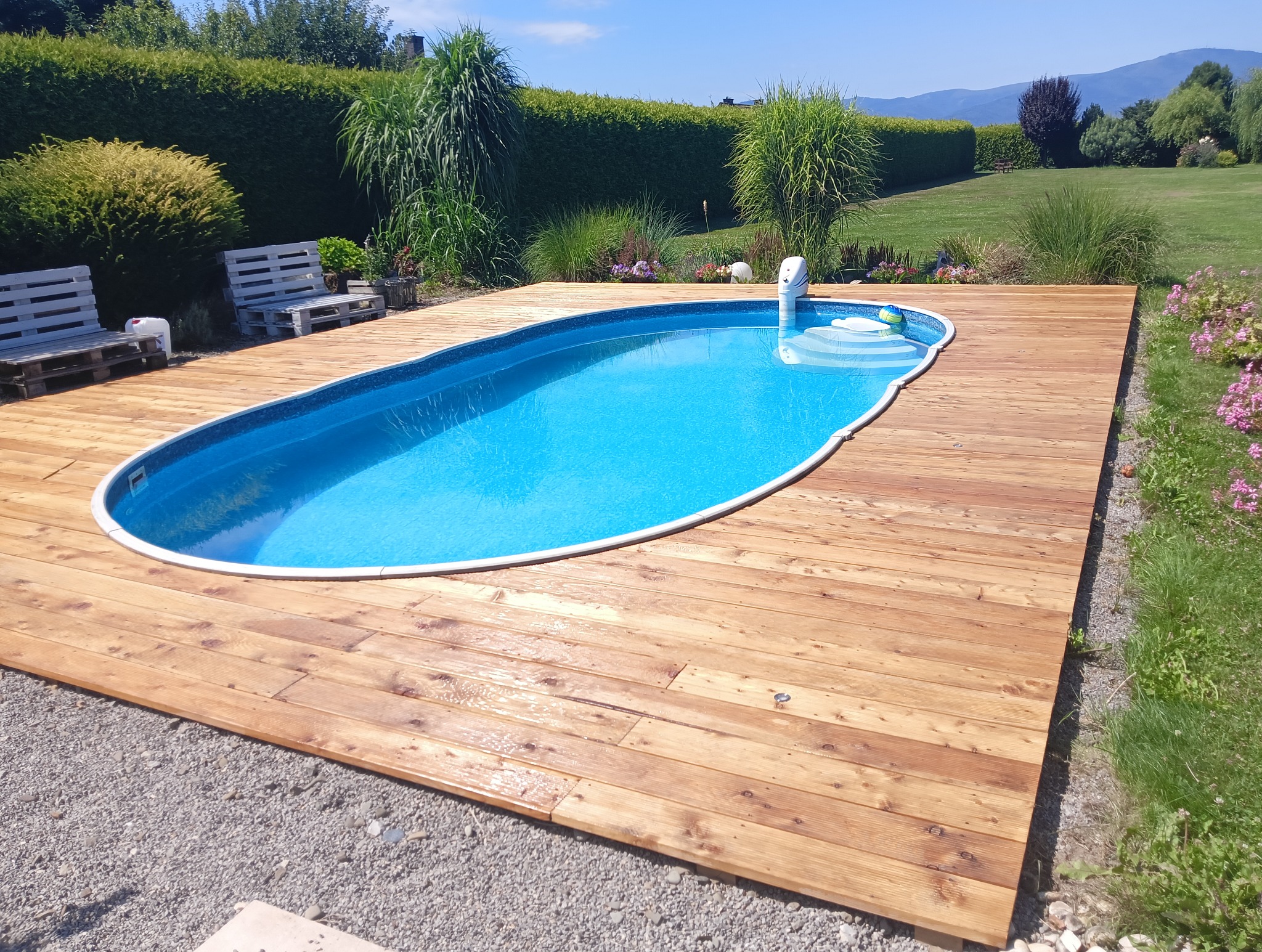 Niebieski basen otoczony drewnianym tarasem, z widokiem na zielony trawnik, ozdobne krzewy i góry w tle.