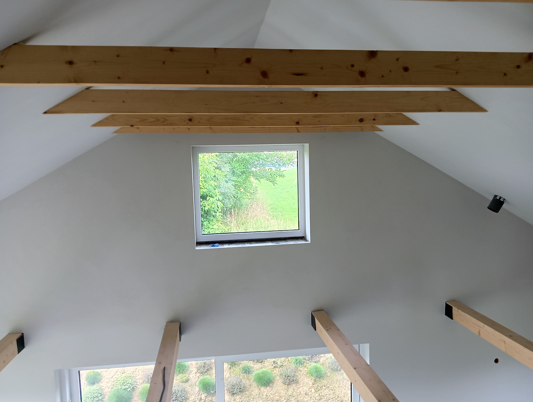Poddasze po remoncie z drewnianą konstrukcją więźby dachowej, pomalowane na biało, z oknem dachowym z widokiem na zieleń.