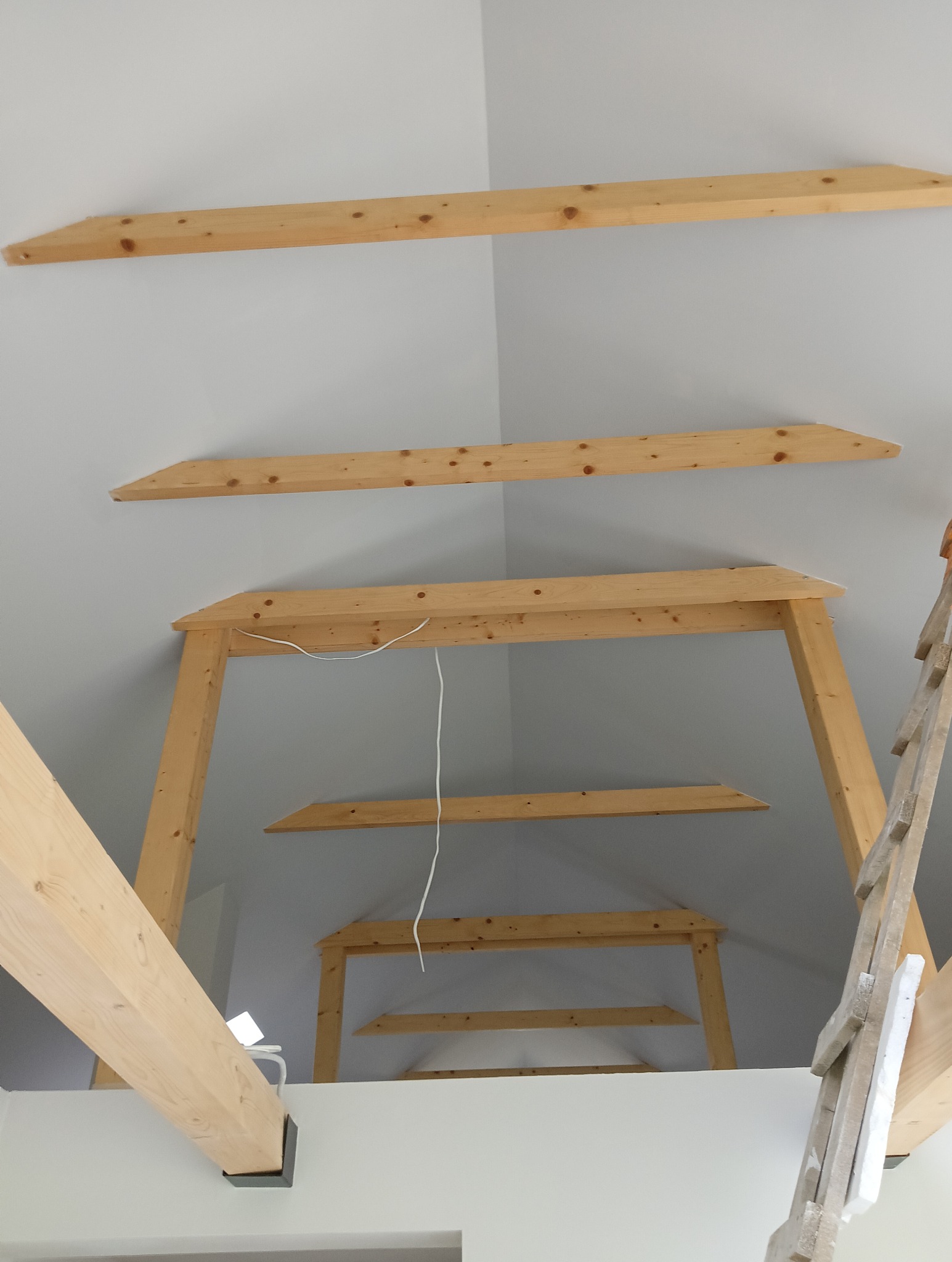 Drewniana konstrukcja więźby dachowej poddasza z widocznymi belkami i elementami mocującymi, częściowo oświetlona, z fragmentem drabiny opierającej się o ścianę.