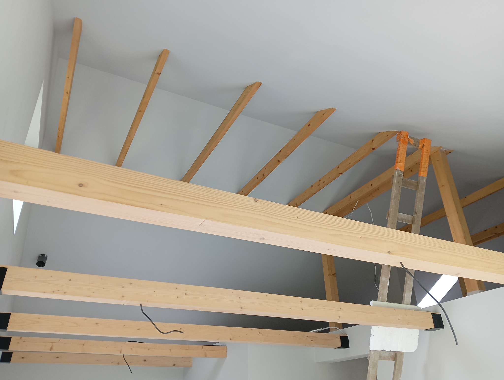 Konstrukcja drewniana poddasza z widocznymi belkami, częściowo ocieplona, z użyciem drabiny zabezpieczonej taśmą, w trakcie prac wykończeniowych.