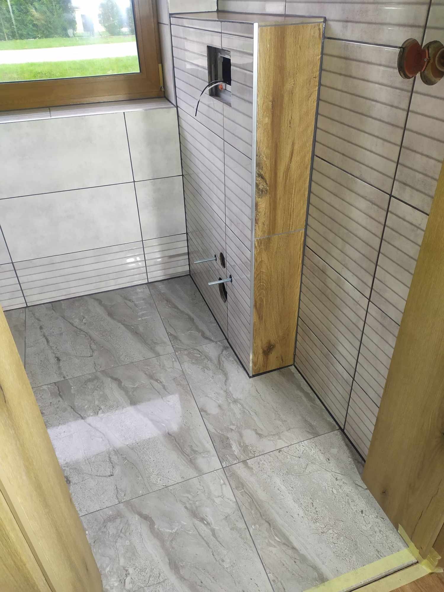 Szare płytki podłogowe imitujące marmur w łazience, obudowa stelaża WC z drewnopodobną okleiną, częściowo wykończona ściana z poziomych płytek w odcieniach szarości, widoczne wyprowadzenia instalacji.