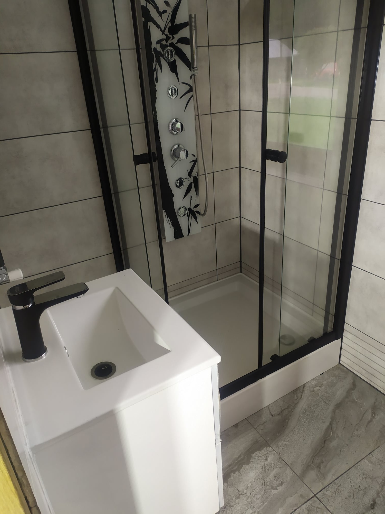 Nowoczesna łazienka z kabiną prysznicową z czarnymi ramami i panelem z motywem bambusa, biała umywalka z czarną baterią, szare płytki podłogowe imitujące marmur.