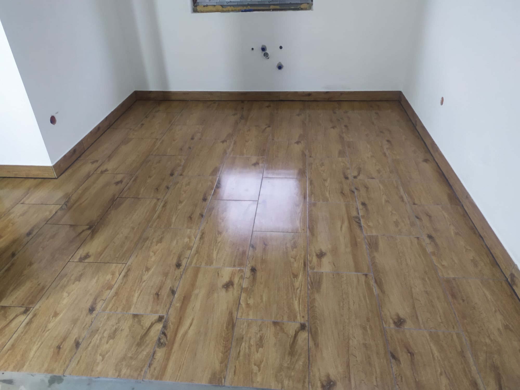 Ułożona podłoga z płytek imitujących drewno w pomieszczeniu z białymi ścianami i widocznymi przyłączami hydraulicznymi.