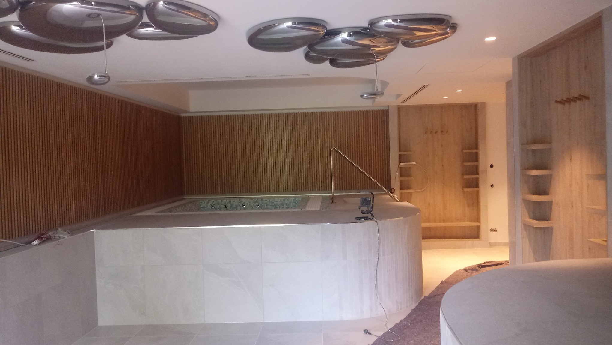 Nowoczesna łazienka z jacuzzi w trakcie wykańczania, widoczne drewniane panele ścienne, okrągłe lustrzane lampy sufitowe i fragment instalacji elektrycznej przy wannie.