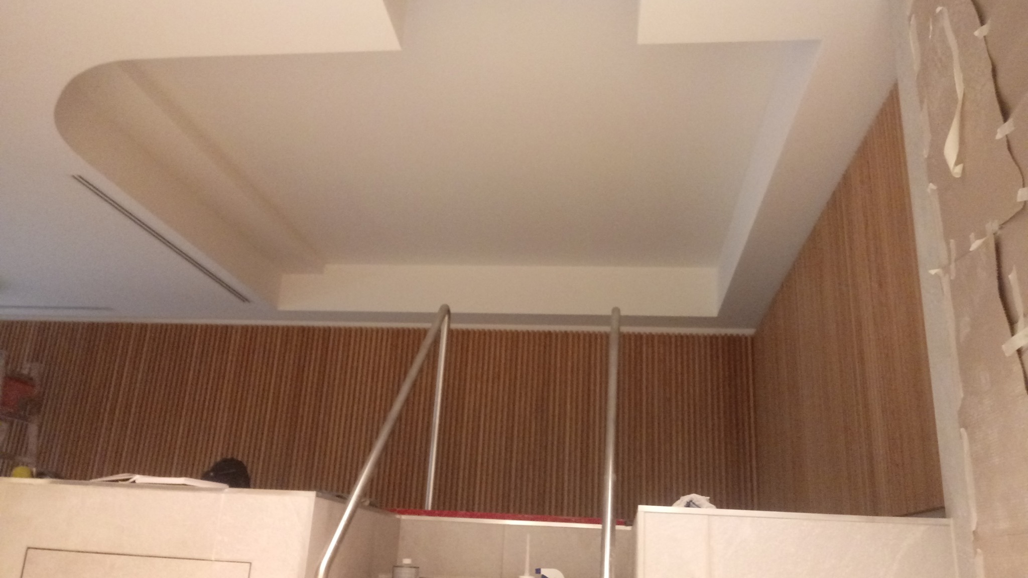 Wykończony sufit podwieszany z oświetleniem liniowym i dekoracyjną listwą, brązowe panele ścienne w tle, fragment metalowej konstrukcji i ściana w trakcie prac wykończeniowych.