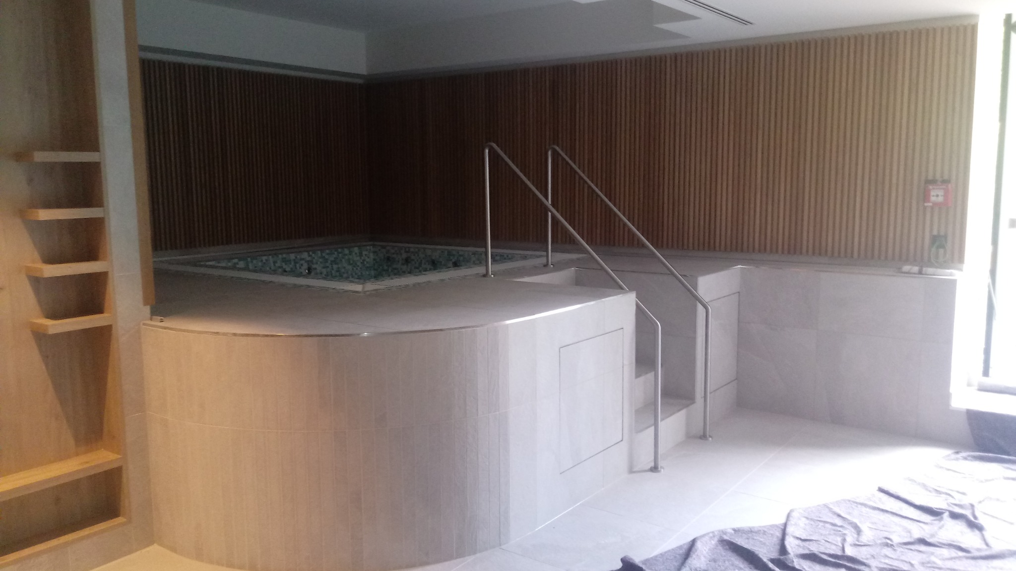 Wnętrze łazienki z jacuzzi wykończone jasnymi płytkami ceramicznymi, drewniane lamele na ścianie, metalowe schodki z poręczą prowadzące do wanny.