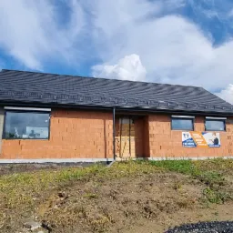 Surowy stan domu jednorodzinnego z czerwonej cegły z czarnym dachem, widoczne okna, reklama firmy na ścianie, obok budynek garażu i biały samochód dostawczy, błękitne niebo z białymi chmurami.