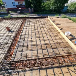 Zbrojenie stropu z siatki stalowej na deskowaniu, widoczne pustaki szalunkowe, narzędzia i częściowo zabudowane elementy konstrukcyjne domu jednorodzinnego w tle.