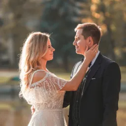 Para młoda w eleganckich strojach, kobieta dotyka twarzy mężczyzny na tle jeziora i zamku w słoneczny dzień.