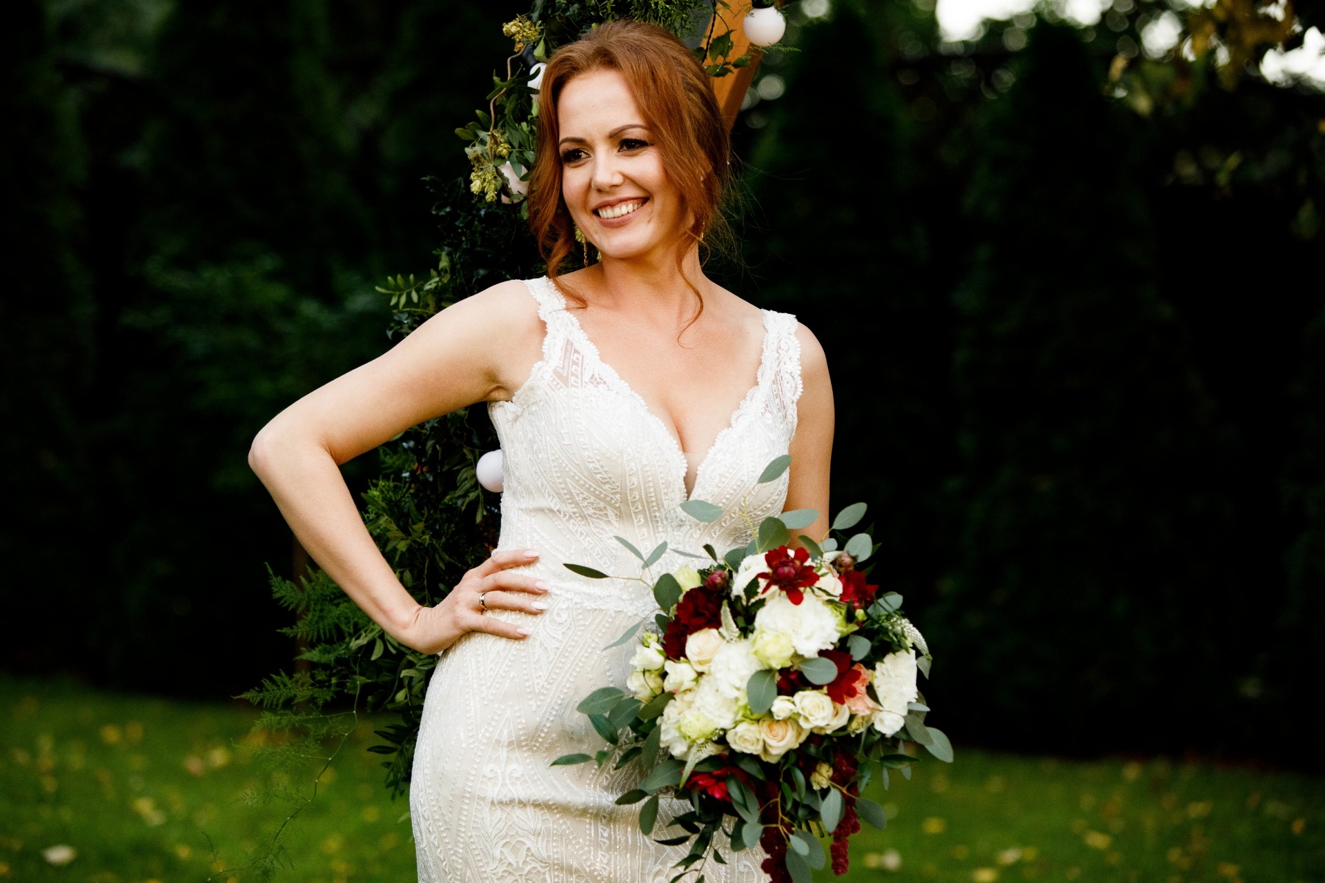Uśmiechnięta panna młoda w koronkowej sukni z bukietem białych i czerwonych róż na tle zieleni.