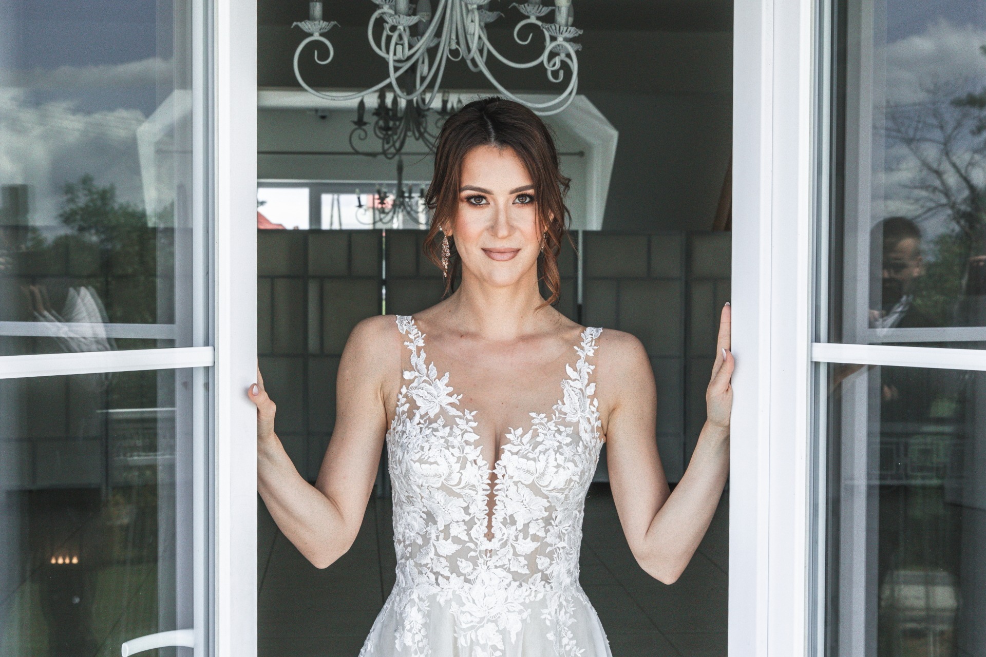 Panna młoda w białej sukni ślubnej z koronkowymi aplikacjami, stojąca w otwartych białych drzwiach z widocznym żyrandolem w tle.
