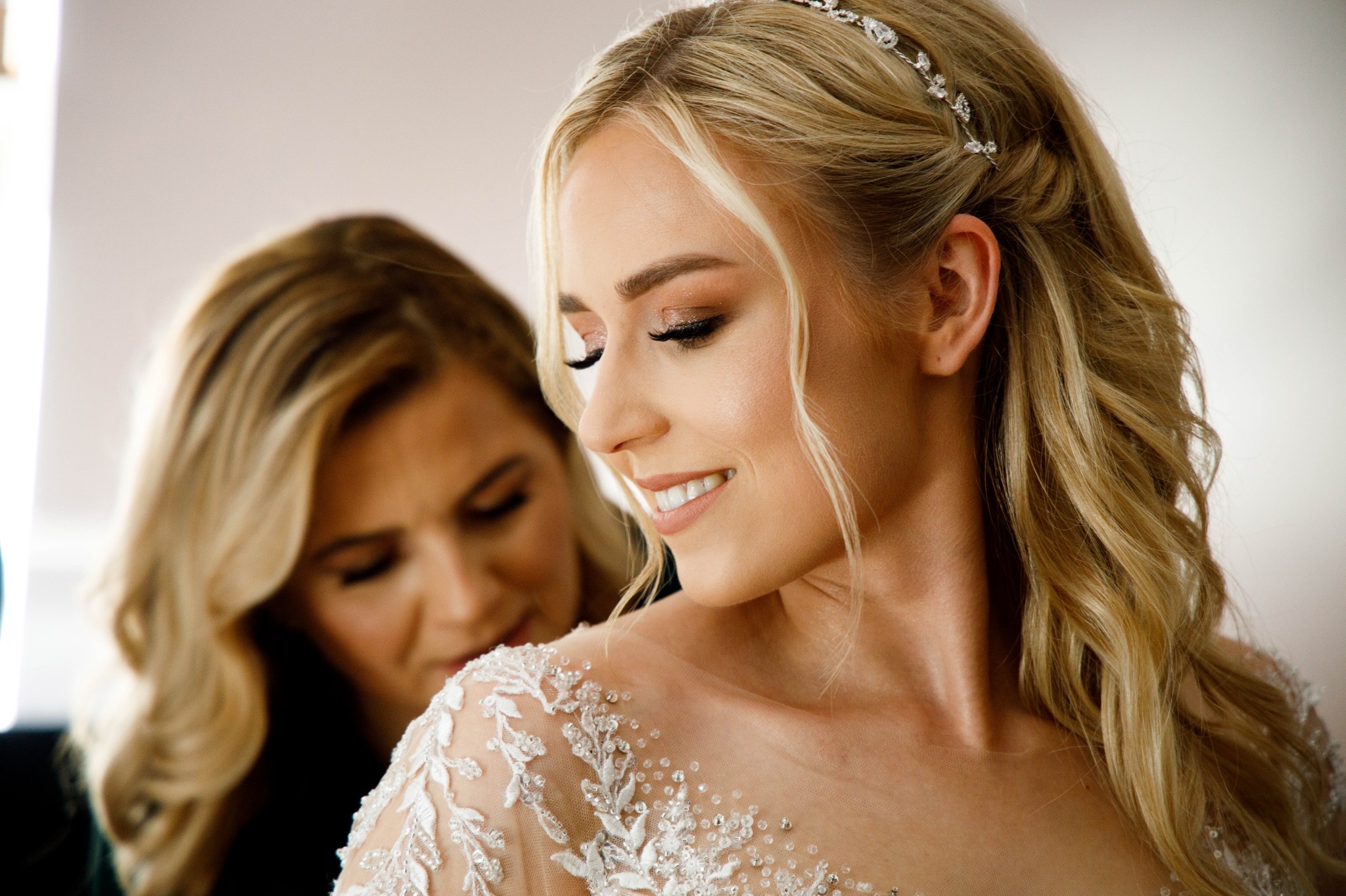 Panna młoda w sukni ślubnej z delikatnymi zdobieniami, uśmiechnięta, z tiarą we włosach, przygotowywana do ślubu przez drugą kobietę.