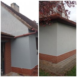PAW-BUD usługi ogólnobudowlane - Fragment domu z szarą elewacją, ceglaną podmurówką i rynną, widoczne detale faktury tynku i łączenia elementów.