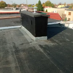 Płaski dach pokryty papą termozgrzewalną z czarnym kominem z metalowym wkładem kominowym, widok z g&oacute;ry na tle zabudowy miejskiej.