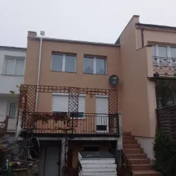 Dwupiętrowy budynek z beżową elewacją, balkonem z kratką i kwiatami, rynnami oraz schodami wejściowymi, widoczny w pochmurny dzień.